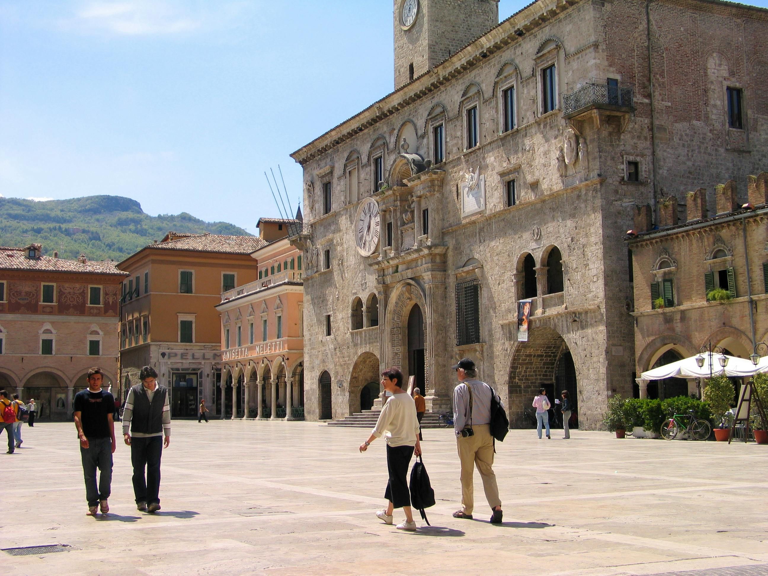 People strolling through a sun-lit Italian piazza beside a historic stone town hall with arched windows and a clock tower