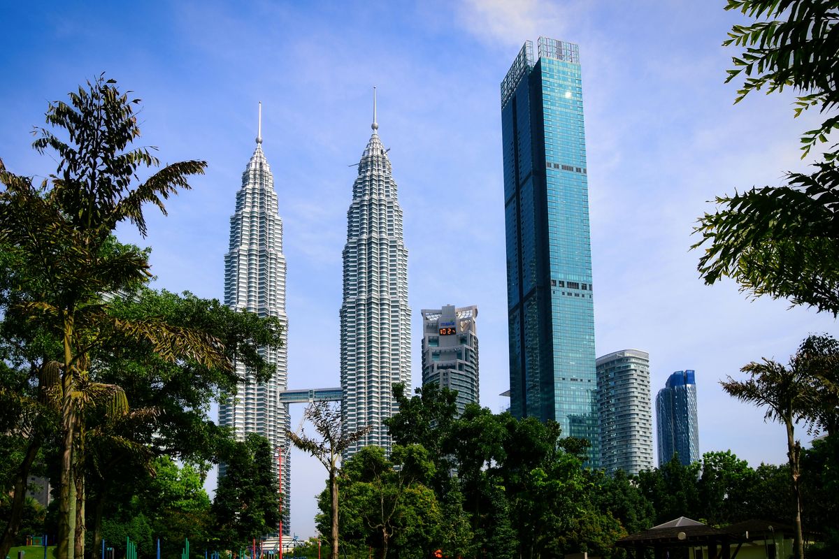 Kuala Lumpur skyline featuring the Petronas Twin Towers framed by lush green trees of KLCC Park under a clear blue sky