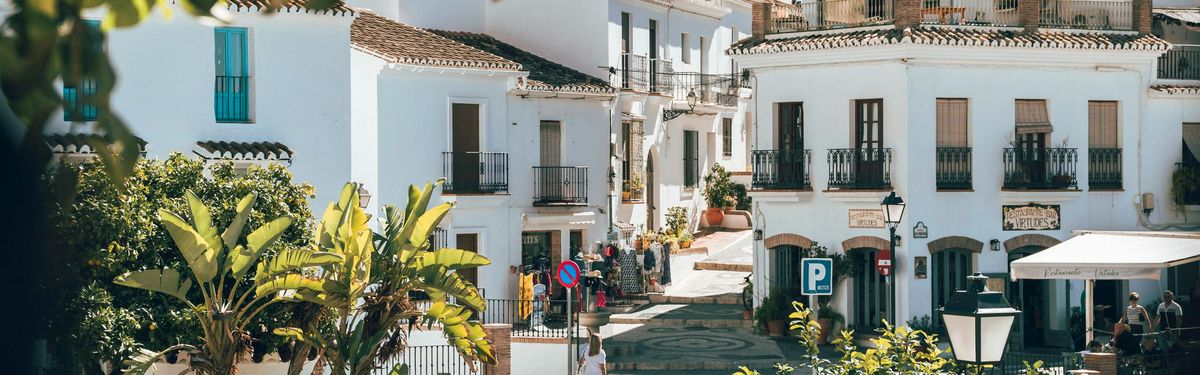 A sunlit cobblestone square in a white-walled Andalusian village with tropical greenery and terrace cafés